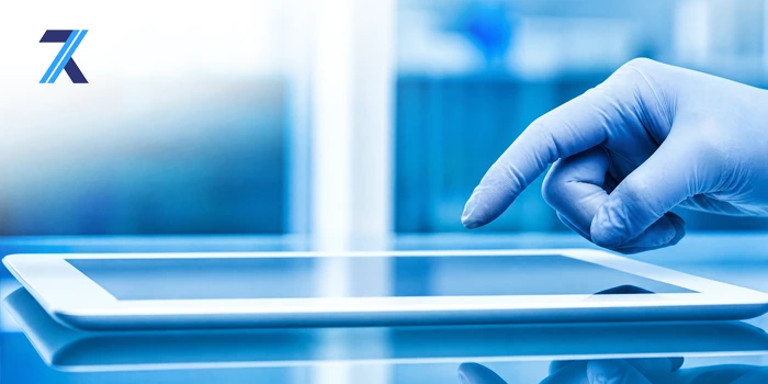 A hand wearing a blue medical glove tapping the screen of a white tablet device on a reflective surface against a blurred blue background with a logo in the top left corner.